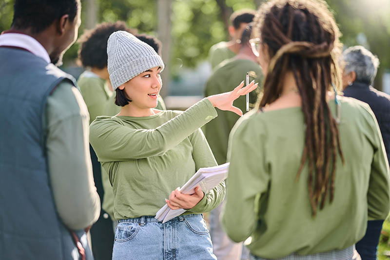 Volunteer schedule, community cleaning leader and charity service outdoor with Asian woman talking. Recycle team, collaboration and eco friendly female manager giving recycling teamwork orders